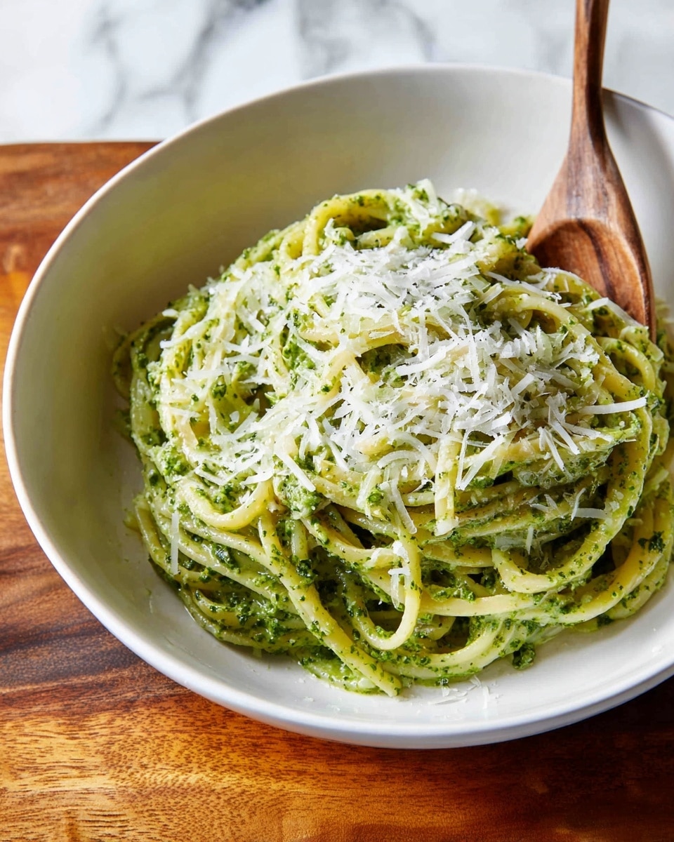 A white bowl holds a neatly twirled pile of long pasta covered in green pesto sauce, creating a thick, textured layer with visible herb specks. On top, there is a light layer of shredded white cheese scattered evenly, adding a soft texture contrast. A wooden spoon is placed inside the bowl, resting against the pasta. The bowl sits on a wooden surface with a white marbled background. Photo taken with an iphone --ar 4:5 --v 7
