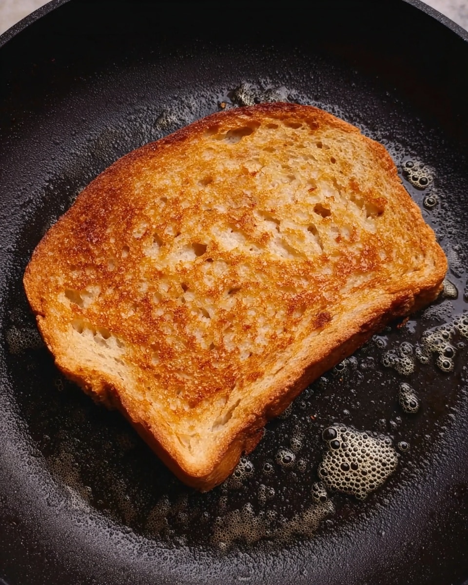 A single slice of golden brown toast is shown in a black frying pan. The toast has a crispy texture with small holes, and the edges are slightly darker than the center, showing it is well toasted. The pan's surface appears oily with small bubbles around the toast. The background is a white marbled texture. photo taken with an iphone --ar 4:5 --v 7