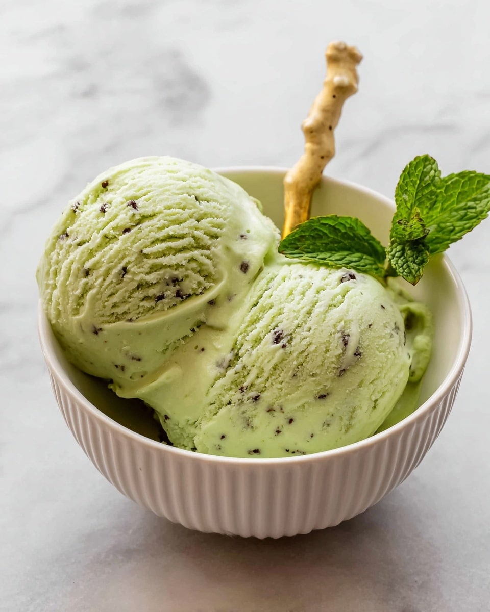 Two big scoops of light green ice cream with tiny dark specks sit closely together in a white bowl with ribbed sides. A gold spoon with a handle shaped like a light beige coral rests inside the bowl behind the ice cream. A small fresh green mint sprig with textured leaves leans against the ice cream on the right side. The bowl is placed on a smooth white marbled surface with soft gray veins. Photo taken with an iphone --ar 4:5 --v 7
