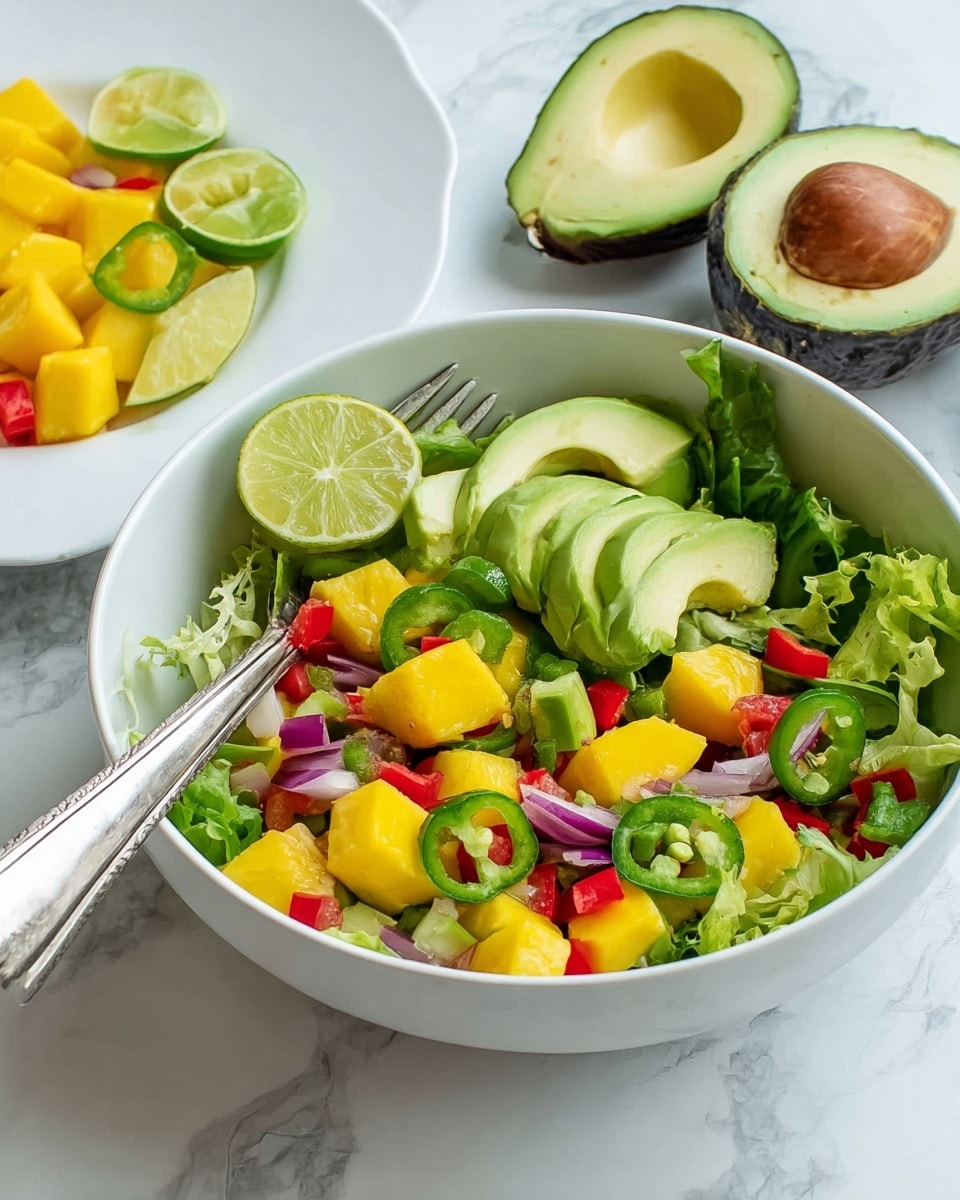A white bowl filled with a colorful salad showing multiple layers: the base layer is fresh green lettuce leaves, topped with chunky cubes of bright yellow mango and pale green avocado slices. Mixed within are slices of green jalapeño, small diced red bell peppers, and thin pieces of purple onion. A half lime with its light green juicy interior is placed on the left side of the bowl. A silver fork rests inside the bowl, leaning on the right edge. In the background, on a white marbled surface, there is another white plate with more mango and avocado pieces, and a halved avocado with a visible brown seed sits to the upper right. The photo taken with an iphone --ar 4:5 --v 7