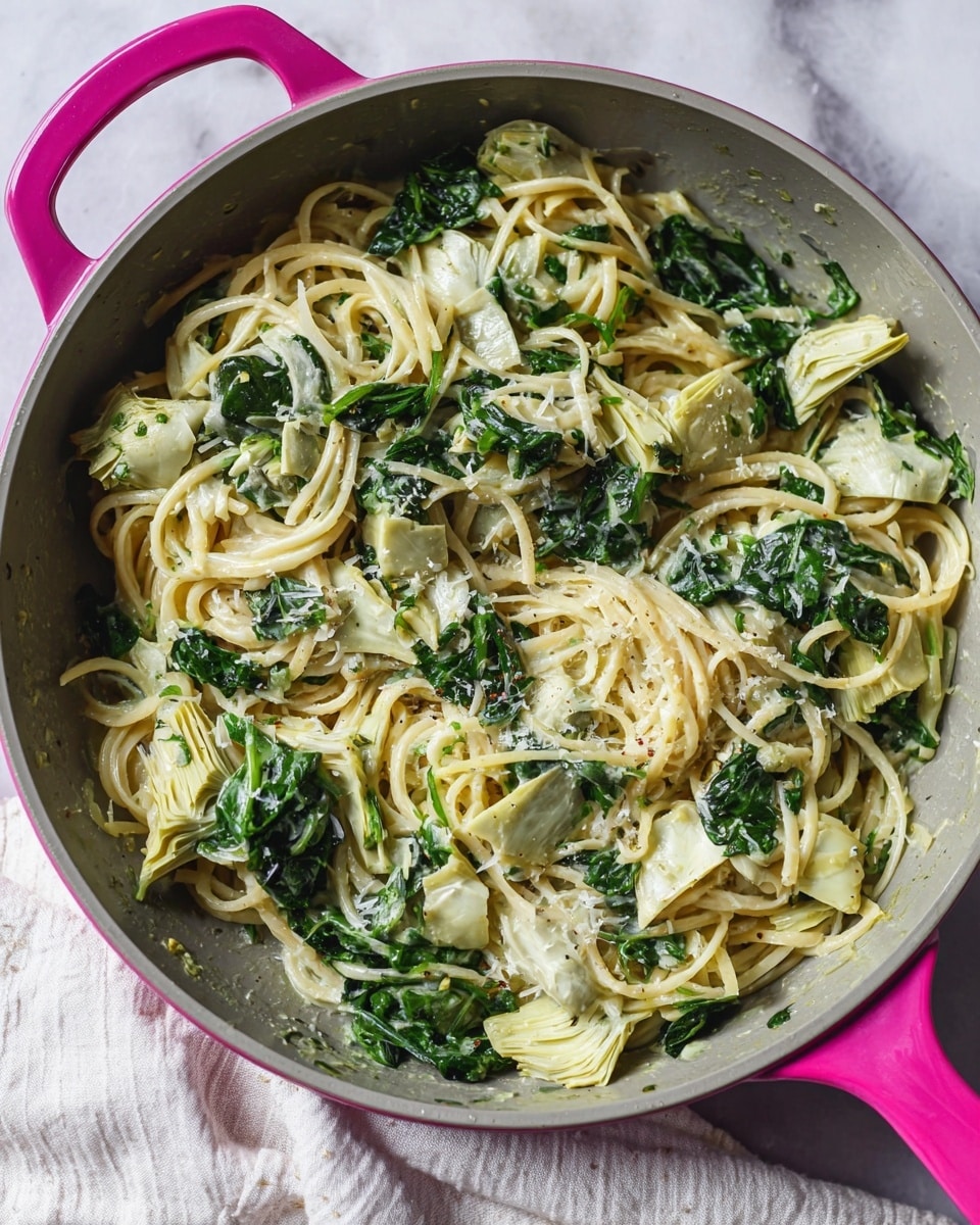 A gray pan with a pink handle holds a pasta dish with one main layer of pale yellow spaghetti mixed with dark green spinach leaves and light green, soft artichoke pieces scattered throughout. The ingredients are mixed evenly, with the spinach and artichoke pieces adding texture and color contrast to the smooth spaghetti. The pan sits on a white marbled surface covered partly by a light fabric. photo taken with an iphone --ar 4:5 --v 7