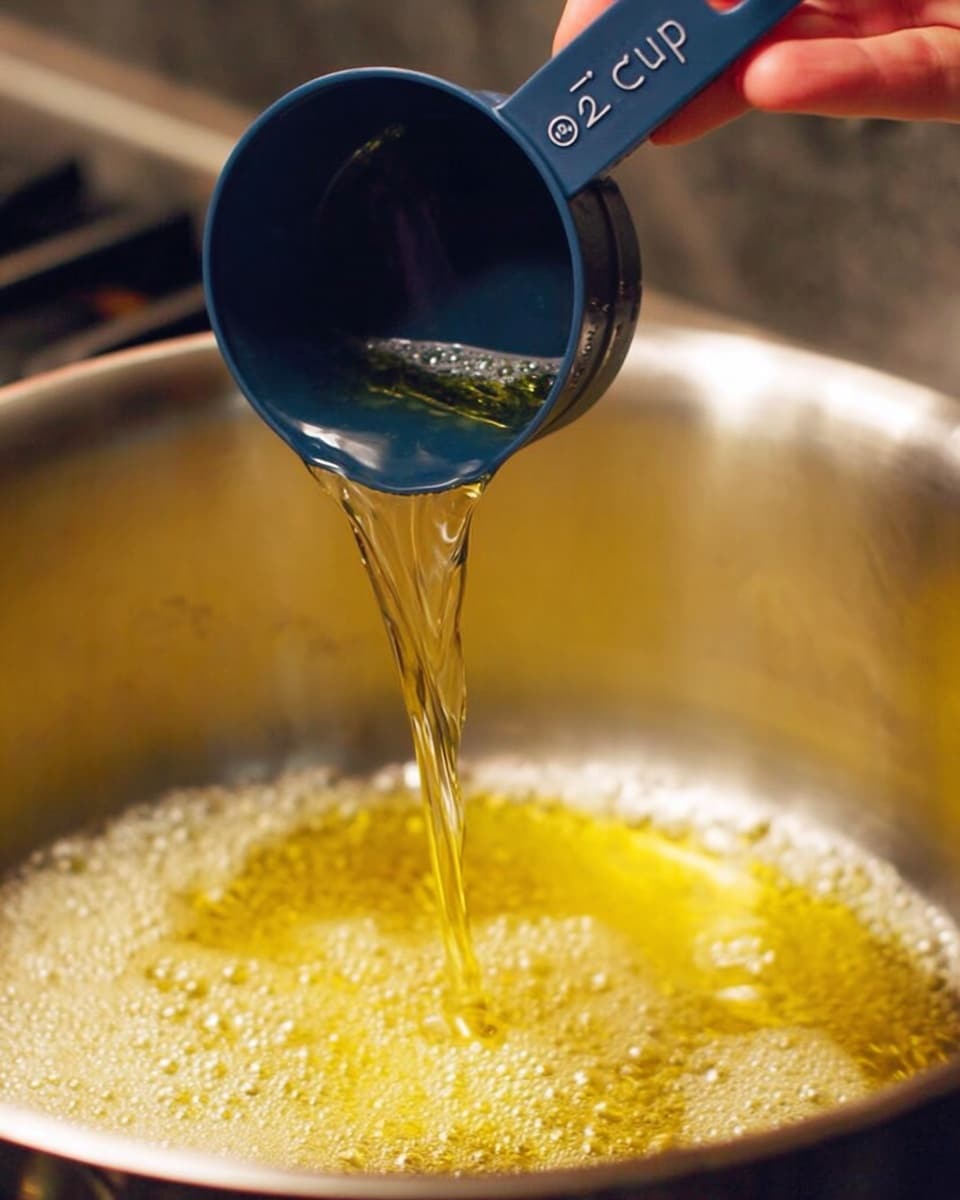 A close-up view of a metal pan with bubbling yellow oil inside, where clear liquid is being poured from a dark blue measuring cup marked