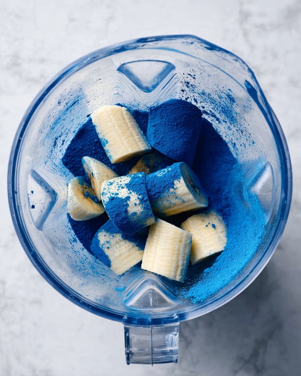 Top view of a clear blender jar on a white marbled surface, containing several pieces of peeled banana covered with a bright blue powder. The bananas are light yellow and soft in texture, scattered irregularly inside the blender. There is a thick layer of intense blue powder coating the bottom and partially covering the bananas, creating a strong contrast between the blue and the pale bananas. Photo taken with an iphone --ar 4:5 --v 7