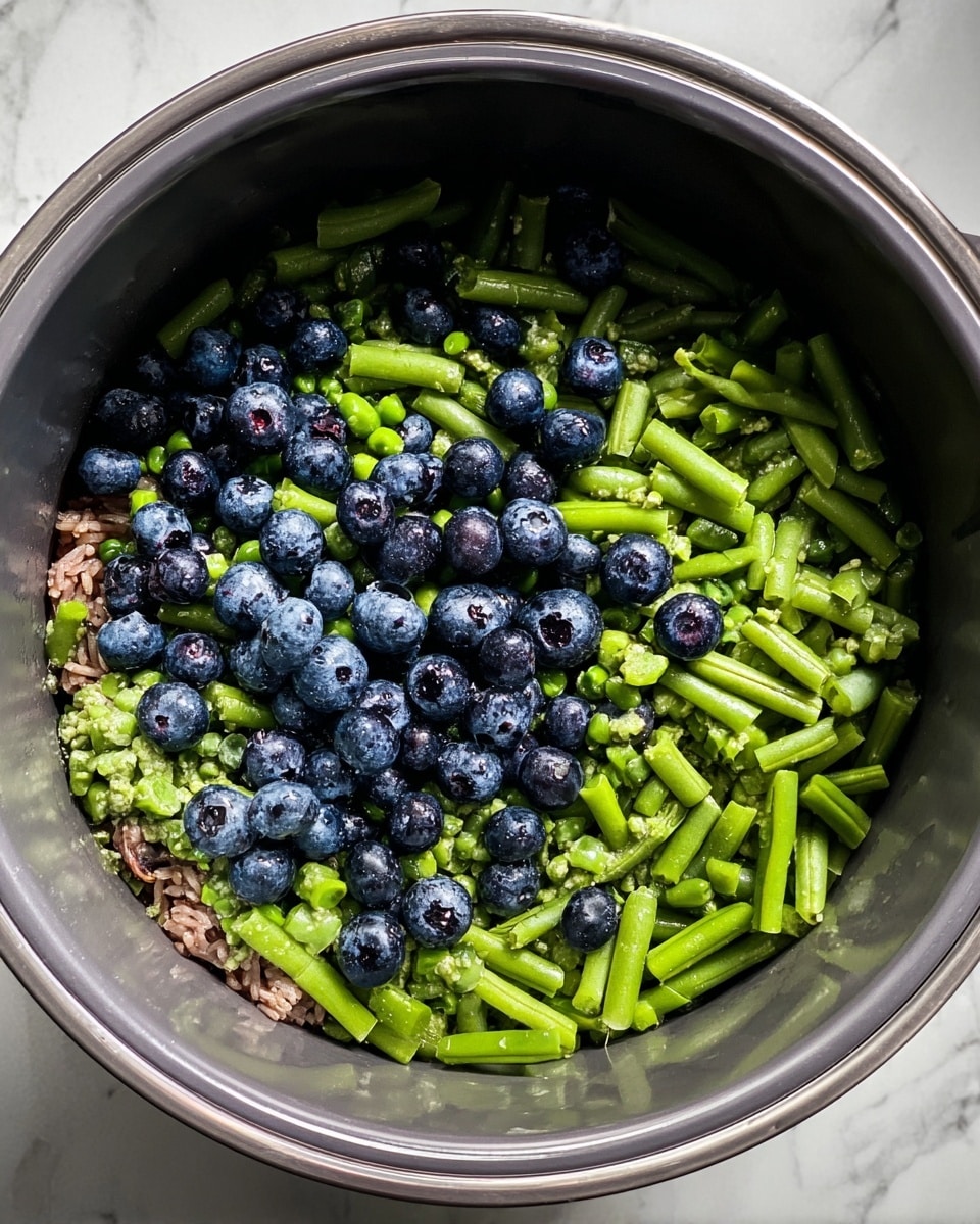 A round gray cooking pot sits on a white marbled surface, filled with three visible layers of ingredients. The bottom layer is not fully shown but has some brown pieces, possibly meat or grains. Above that, green peas and green bean pieces form the middle and top layers, with the green beans cut into small segments and scattered throughout. Dark blue blueberries are mixed evenly on the top layer, contrasting with the fresh green colors of the vegetables. The pot’s smooth inner surface has a metallic sheen, and the whole scene has natural lighting, showing clear colors and textures. Photo taken with an iphone --ar 4:5 --v 7