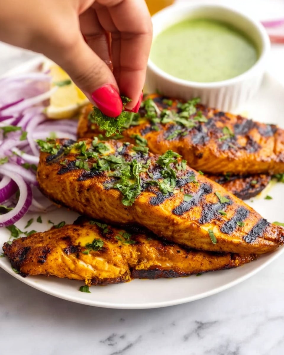 The image shows three grilled fish fillets stacked on a white plate, each fillet with dark grill marks and a bright orange-brown color from the spices. The top fillet is being garnished with fresh green herbs by a woman's hand with red-painted nails. Around the plate are slices of purple onion and a small round white bowl filled with a green sauce. The whole scene is set on a white marbled surface. photo taken with an iphone --ar 4:5 --v 7