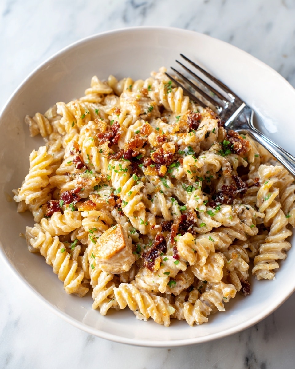 The dish shows a white plate filled with rotini pasta mixed with a creamy sauce, light beige in color. Scattered on top and mixed within the pasta are small, dark reddish-brown crispy bits. There are pieces of cooked chicken with a light beige color and some specks of herbs visible. Small green herbs are sprinkled over the pasta, adding a touch of color. A fork with a silver head and black handle rests on the top right edge of the plate, which is placed on a white marbled surface. Photo taken with an iphone --ar 4:5 --v 7