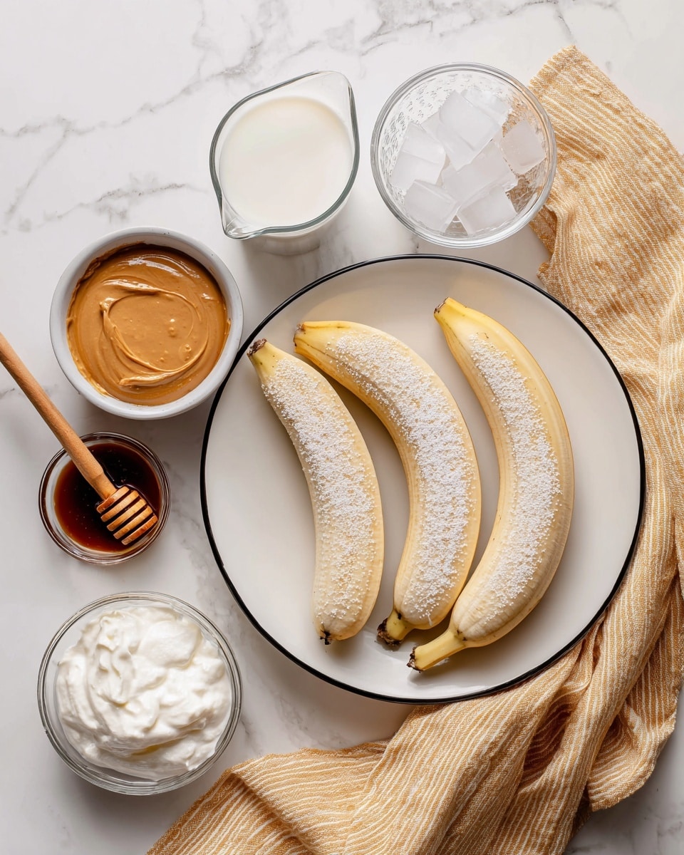 The image shows four peeled bananas on a white plate with a thin black rim, each banana lightly dusted with a powdery texture. To the left, there is a small white bowl filled with smooth peanut butter, a clear glass measuring cup with white milk, and a small white bowl with dark honey and a wooden honey dipper resting inside. Below these ingredients is a small glass bowl with white whipped cream and a larger glass bowl filled with clean ice cubes. A soft mustard-yellow cloth with white stripes is draped loosely beneath some of the bowls, all set against a white marbled surface. Photo taken with an iphone --ar 4:5 --v 7