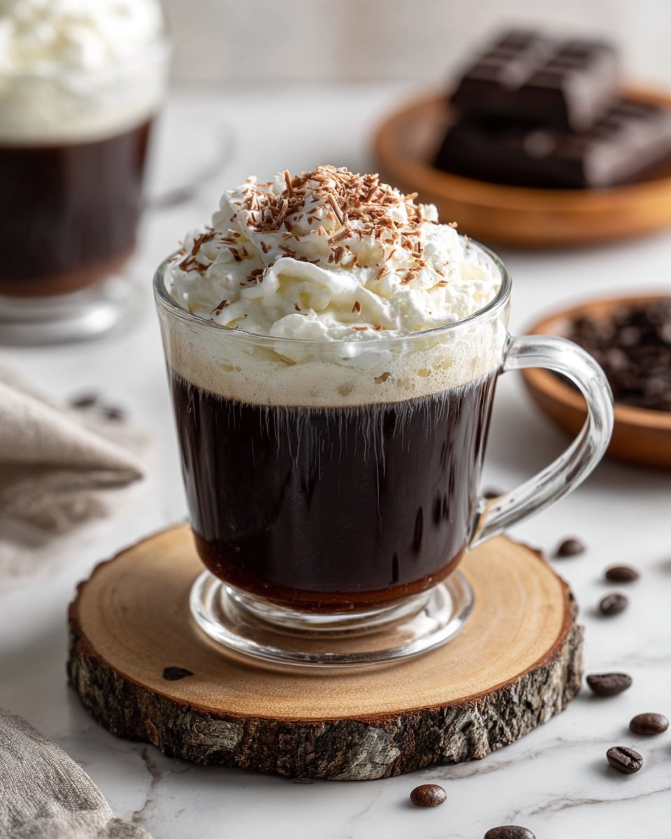 A clear glass mug filled with dark brown coffee forms the base layer, topped generously with a thick layer of soft white whipped cream, sprinkled with fine chocolate shavings that create a light brown texture on top. The mug sits on a round, rustic wooden coaster, which adds a natural, textured contrast to the smooth surfaces. Scattered dark coffee beans lie on the white marbled surface around the mug, adding detail to the setting. In the background, another similar mug with whipped cream and a wooden coaster is partly visible, along with a wooden plate holding chocolate pieces. Photo taken with an iphone --ar 4:5 --v 7