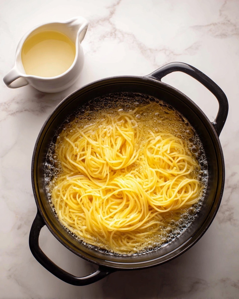 A black cast iron pot filled with cooked yellow spaghetti noodles submerged in clear, steaming water with small bubbles on the surface. To the upper left of the pot, there is a small white pitcher holding a pale liquid. Both items are placed on a white marbled surface. photo taken with an iphone --ar 4:5 --v 7