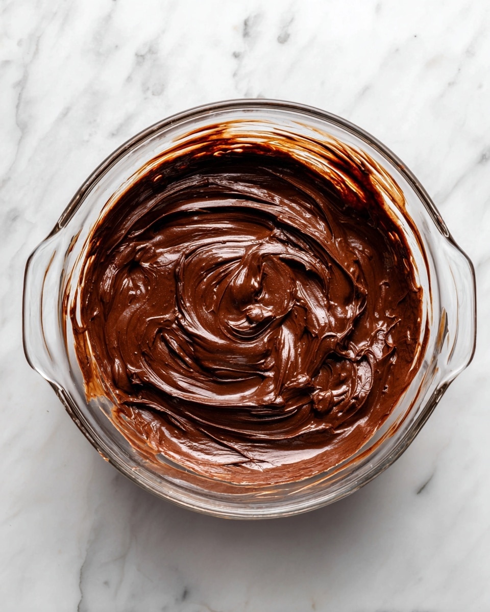 A clear glass bowl sits on a white marbled surface, holding a thick, rich dark brown chocolate mixture. The chocolate batter covers the bottom and partially swirls up the sides of the bowl, showing smooth, creamy textures with some streaks around the edges. The bowl has two small handles on opposite sides, and the light highlights the glossy finish of the chocolate. Photo taken with an iphone --ar 4:5 --v 7