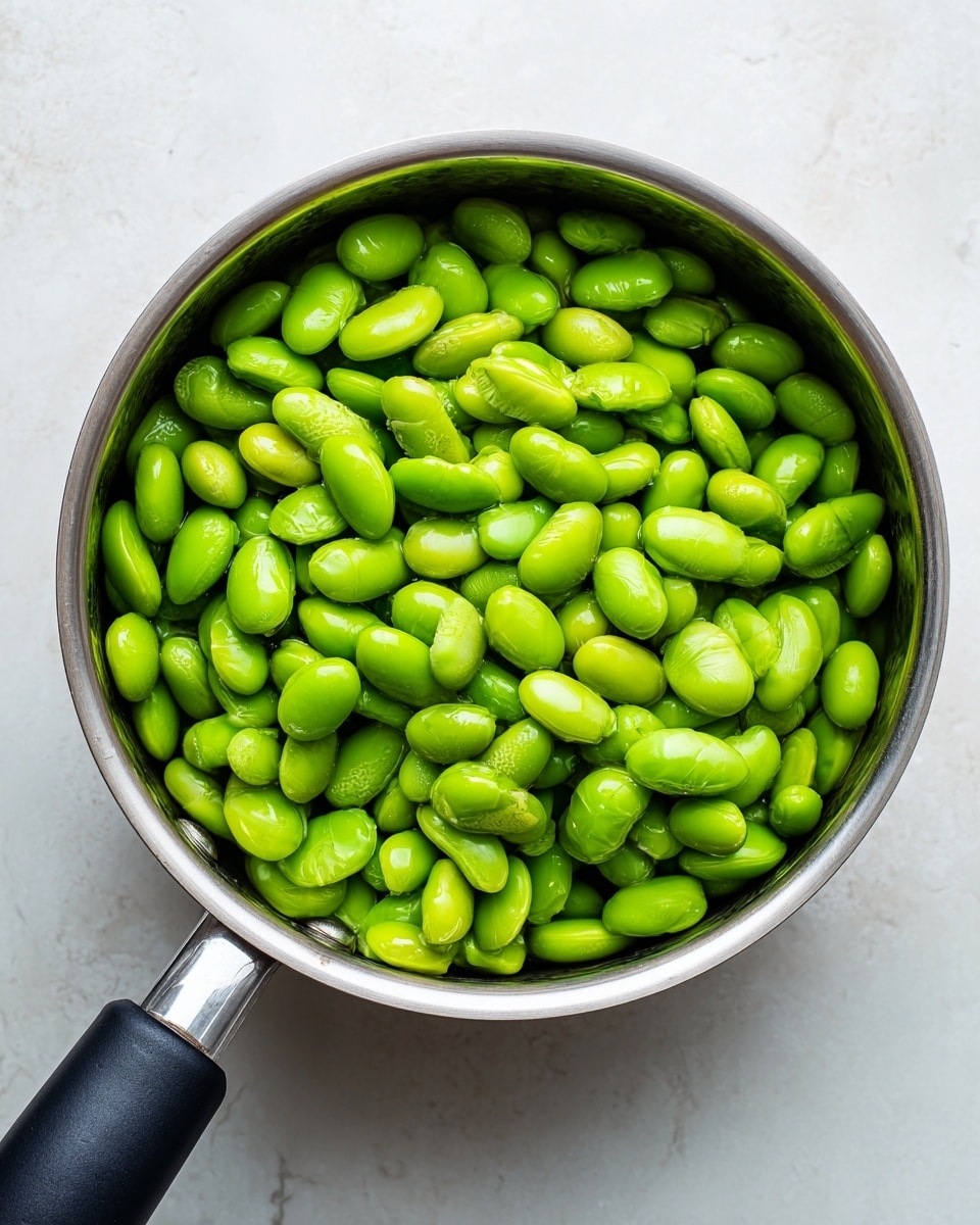 A metal saucepan filled with bright green edamame beans fills the frame from above, sitting on a white marbled surface. The smooth, shiny beans are tightly packed and have a fresh, moist look. The saucepan handle, black with a silver band, extends from the left side, with the clean silver pot rim visible. The background is simple, highlighting the vivid green beans and the metal pot. photo taken with an iphone --ar 4:5 --v 7