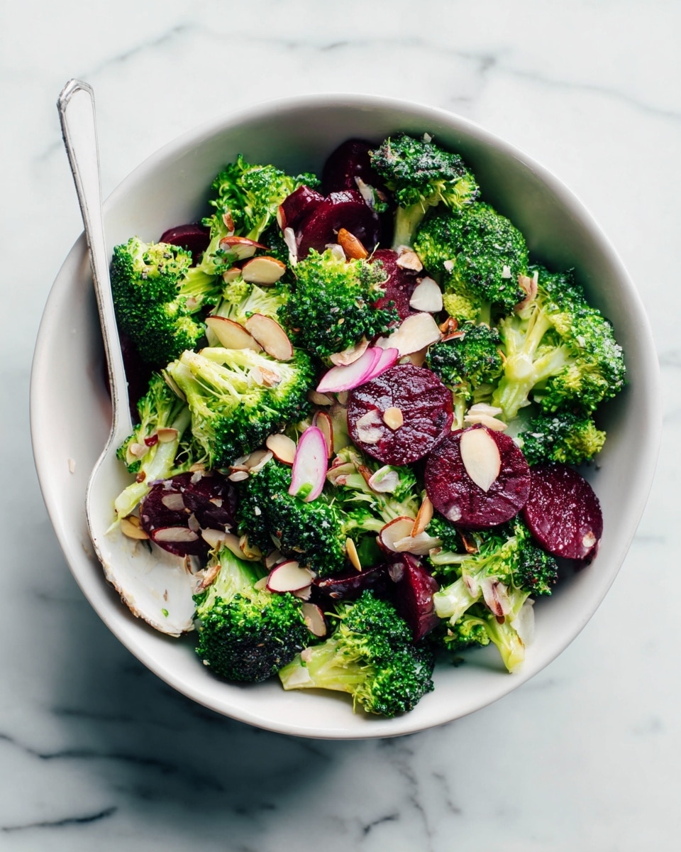 The image shows a white bowl filled with a fresh broccoli salad. The salad has two main layers: bright green broccoli florets with a rough texture and dark red, round beet slices that have a smooth surface. Thin, pale slices of almond and small bits of light pink onions are scattered on top, creating a mix of colors and textures. A white spoon is placed inside the bowl, partly covered by the salad. The bowl sits on a white marbled surface. photo taken with an iphone --ar 4:5 --v 7