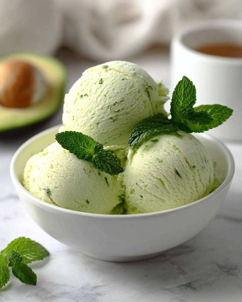 The image shows three light green ice cream scoops arranged closely in a white bowl. The ice cream has a smooth texture with visible small green specks, suggesting a fresh flavor. On top of the scoops, there are three bright green fresh mint leaves adding a vibrant contrast. The bowl rests on a surface with a white marbled texture, and in the blurred background, a halved avocado and a white cup are faintly visible, providing a soft contrast to the focused ice cream. photo taken with an iphone --ar 4:5 --v 7