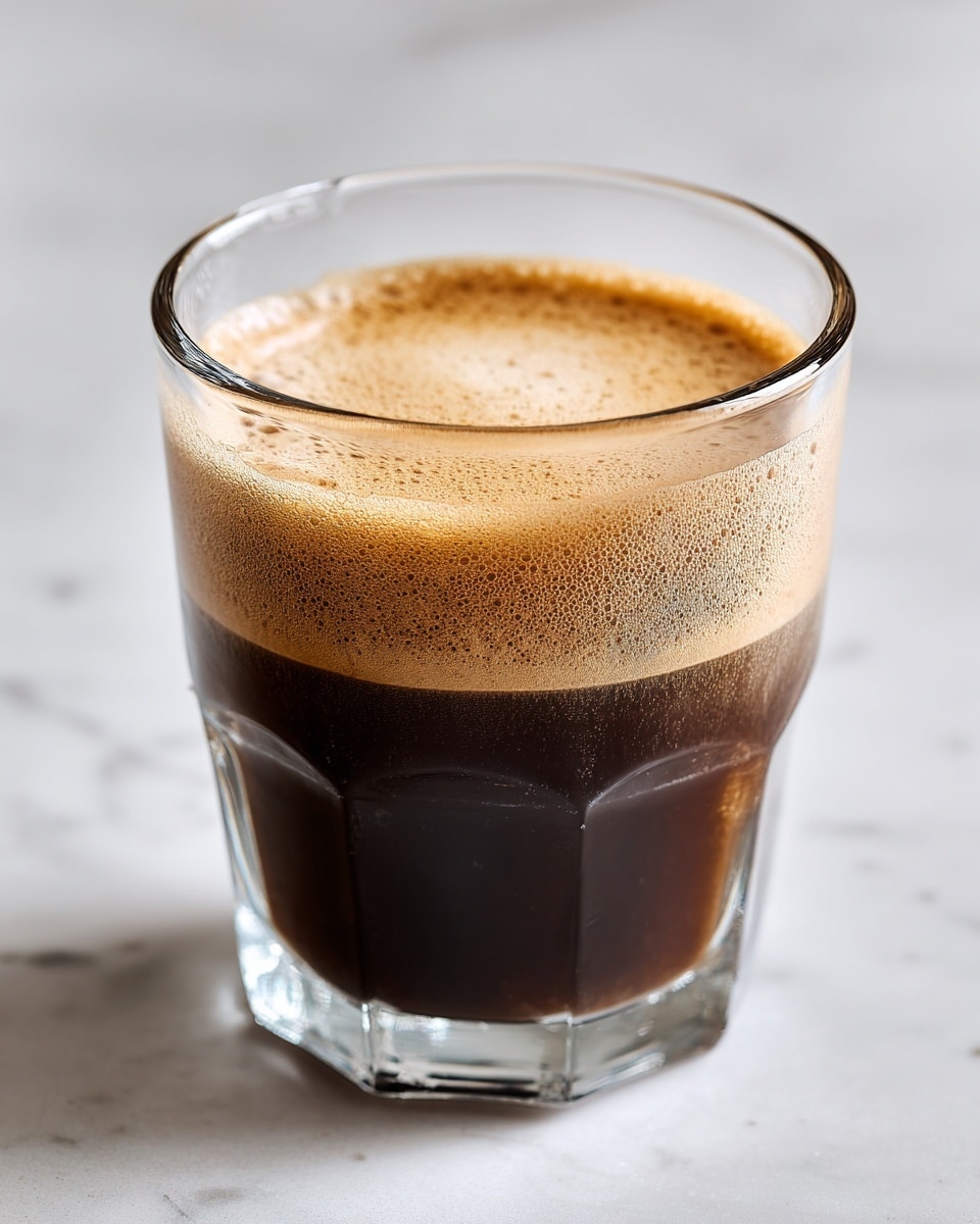 A clear glass filled about halfway with a dark brown coffee liquid has a light tan foam layer, about one-third inch thick, on top. The glass is placed on a white marbled surface with soft lighting and a simple, light gray background. Photo taken with an iphone --ar 4:5 --v 7
