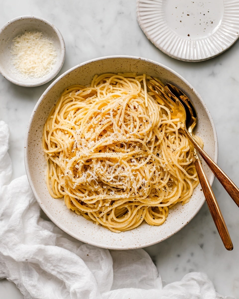 A large white speckled bowl holds a single layer of spaghetti noodles. The noodles are light brown and slightly shiny from sauce, with specks of black pepper and white cheese bits spread across the top. Inside the bowl, there is a pair of vintage-style golden serving utensils with wooden handles resting on the right side, partially tangled in the pasta. The bowl sits on a white marbled surface with a white cloth napkin casually placed underneath the lower left side. Nearby, there are small white bowls with grated cheese and an empty white ridged plate, both set on the same white marbled texture. photo taken with an iphone --ar 4:5 --v 7
