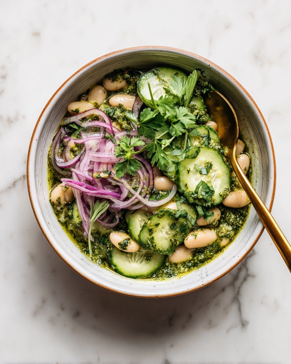 A white bowl with brown rim contains a fresh salad layered with green pesto sauce as the base, topped with white beans submerged in the sauce, thin slices of purple onion scattered on the beans, and thin green cucumber slices arranged on top with fresh green herbs like cilantro and parsley on the very top. A gold spoon rests inside the bowl on the right side. The bowl is placed on a white marbled surface. Photo taken with an iphone --ar 4:5 --v 7