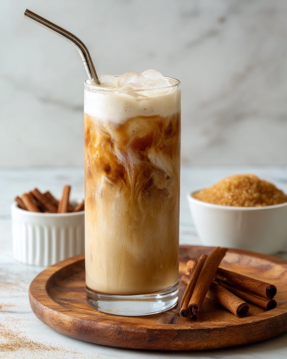 The image shows a tall clear glass filled with a creamy iced coffee drink, with a base layer of light brown coffee and milk swirling inside, topped with a thick, frothy white foam layer. A shiny metal straw is placed inside the glass, leaning toward the left. The glass sits on a round wooden tray with some cinnamon sticks and a small white bowl filled with brown sugar in the background. The surface beneath is a white marbled texture. Photo taken with an iphone --ar 4:5 --v 7