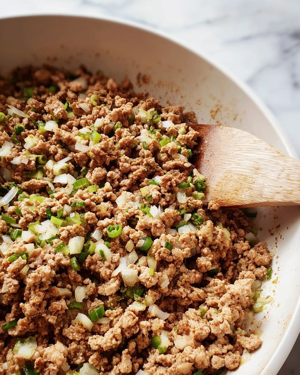 A close-up view of cooked ground meat mixed with small chopped green and white pieces, likely onions or similar vegetables, all inside a white pan with a wooden spoon partially visible on the right side. The ground meat is light brown and crumbly, with a rough texture spread across the pan. The background shows a white marbled surface. photo taken with an iphone --ar 4:5 --v 7