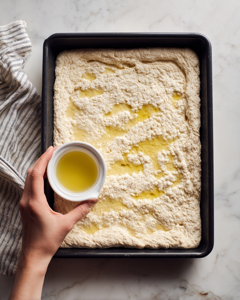 A rectangular piece of dough is spread flat inside a black baking tray, with edges slightly pressed down to form a border. The dough has a rough texture and a pale beige color. A woman's hand is seen holding a small white ramekin and pouring a yellowish oil or butter over the dough in uneven patches near the top and middle. The tray is placed on a white marbled surface, and a striped cloth is partly visible to the left. Photo taken with an iphone --ar 4:5 --v 7
