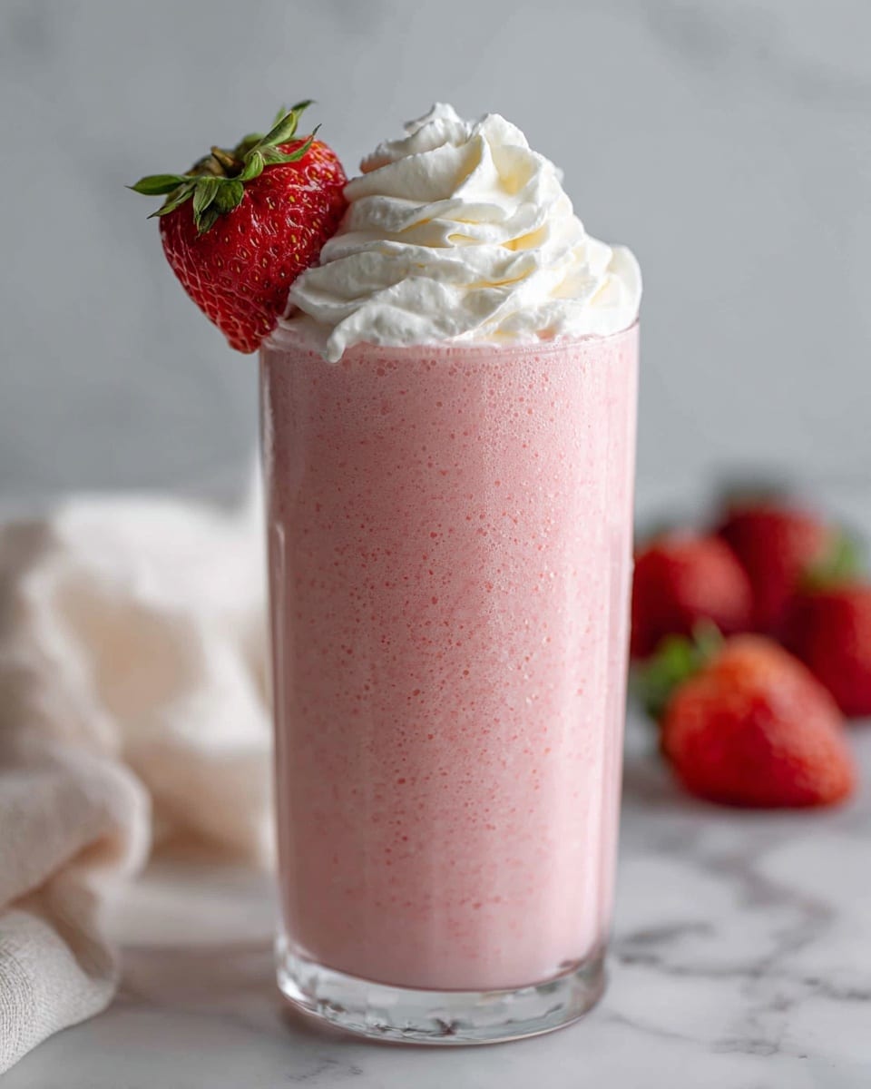 The image shows a tall clear glass filled with a pink strawberry smoothie that has a creamy texture. The smoothie takes up most of the glass and is topped with a thick, white swirl of whipped cream. On the rim of the glass, there is one bright red strawberry with its green leafy top still attached. The glass is placed on a white marbled surface with a soft white cloth nearby and some blurred strawberries in the background. photo taken with an iphone --ar 4:5 --v 7
