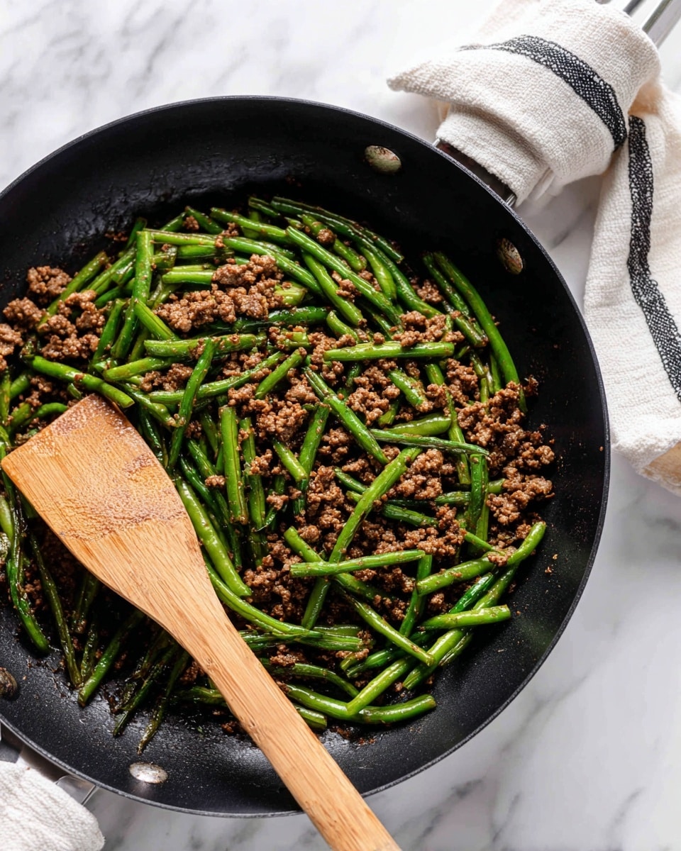 A black frying pan filled with cooked ground meat mixed evenly with bright green long beans, showing a slightly crispy texture on the beans and crumbled meat. A wooden spatula is scooping some of the mixture on the left side of the pan, and the pan handle is wrapped with a white cloth that has black stripes. The pan sits on a white marbled surface, giving a clean and fresh background. photo taken with an iphone --ar 4:5 --v 7
