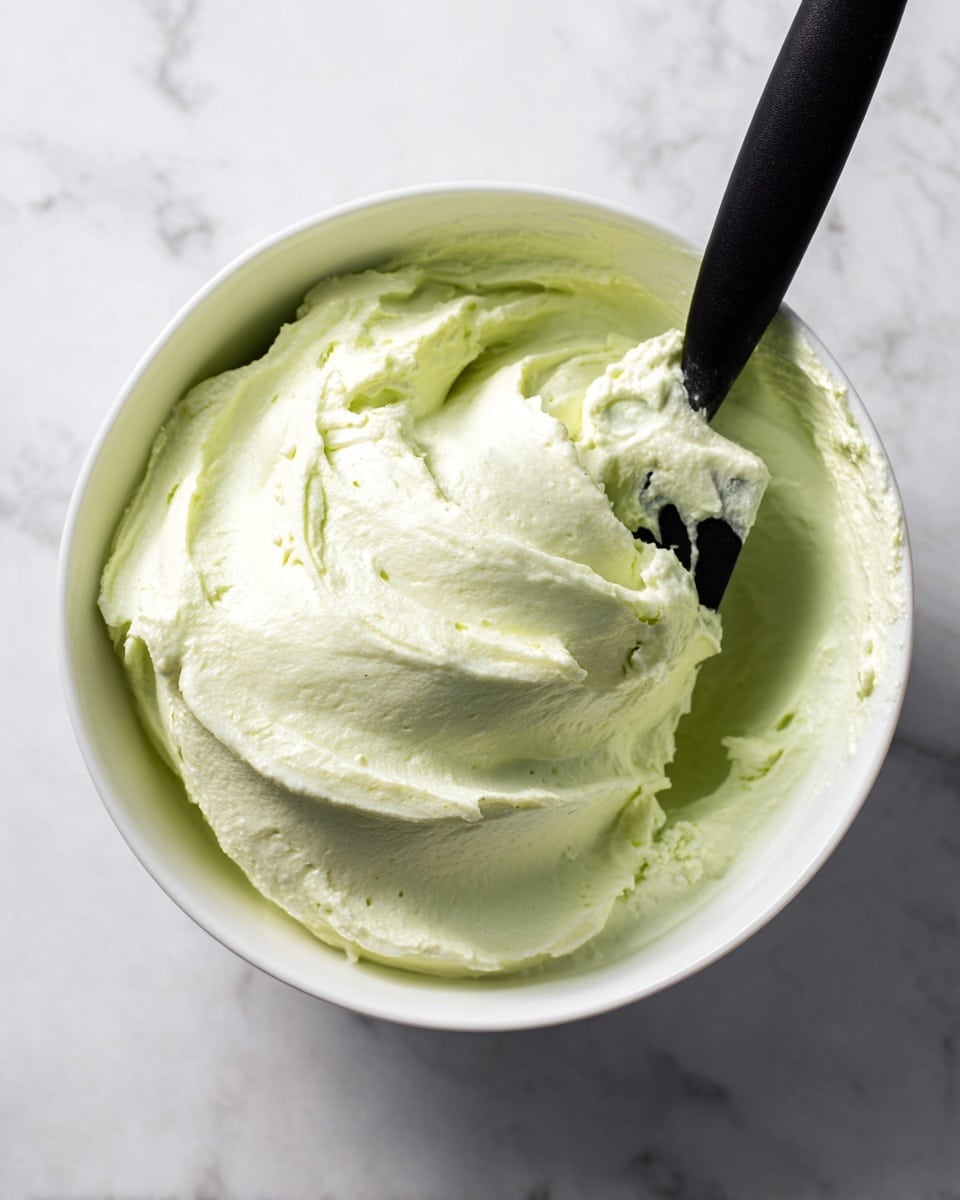 A close-up view of a white bowl filled with smooth, light green cream. The texture is soft and fluffy, with gentle swirls visible on the surface. A black spatula is partially dipped into the cream on the right side of the bowl, with some cream sticking to its edge. The bowl is placed on a white marbled surface. photo taken with an iphone --ar 4:5 --v 7