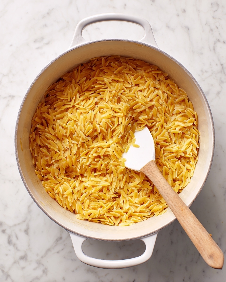 A white pot with two handles contains a single layer of golden yellow orzo pasta being stirred by a white spatula with a light wooden handle, the pasta is spread unevenly across the pot's base showing some surface in between, and the pot is placed on a white marbled texture surface photo taken with an iphone --ar 4:5 --v 7
