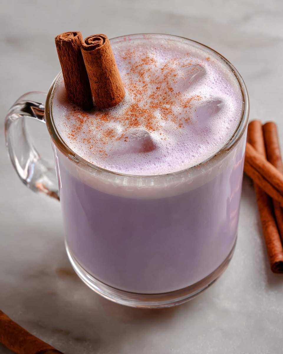 A clear glass mug filled with a light purple frothy drink with ice cubes visible under the foam, topped with two brown cinnamon sticks standing upright and a light sprinkle of brown cinnamon powder on top, placed on a white marbled surface. photo taken with an iphone --ar 4:5 --v 7