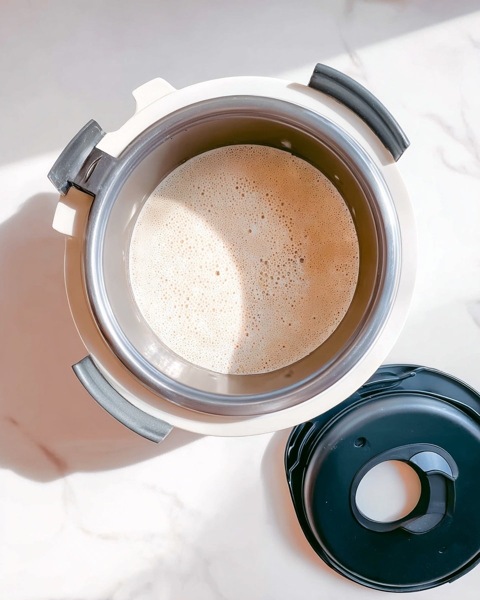 The image shows a top view of a white kitchen appliance filled with a light beige liquid with small bubbles on the surface. The container inside has a metallic look with four small notches around its edge. To the right of the appliance is a black lid with a round handle in the center, placed on the white marbled surface. The appliance handle at the top is metallic silver, and the two side handles of the container are black. The overall setting is bright with soft shadows. Photo taken with an iphone --ar 4:5 --v 7