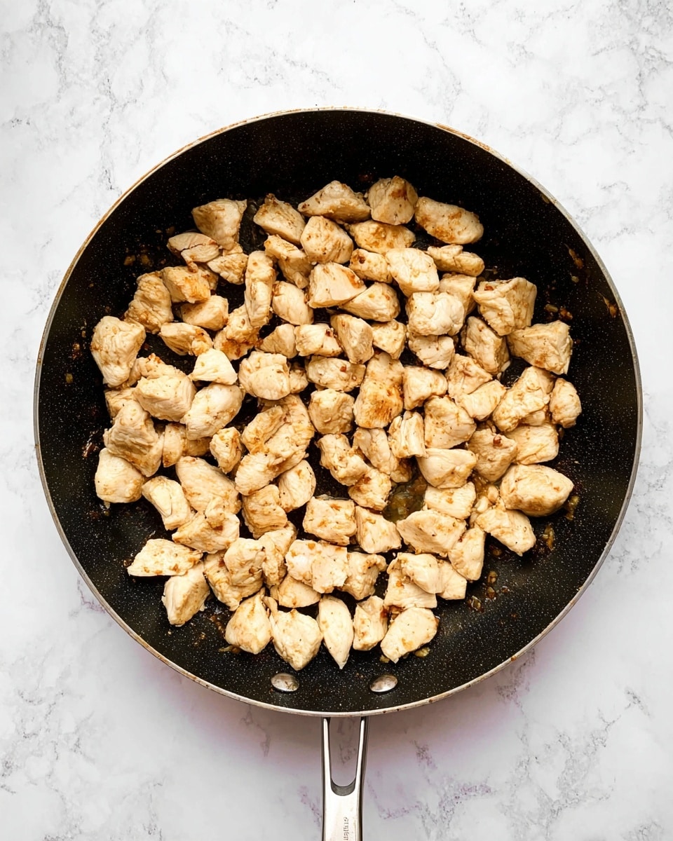 A black frying pan filled with many small, light golden pieces of cooked chicken, evenly spread across the pan surface, showing slight browned spots on the surface of the chicken chunks, all on a white marbled textured background photo taken with an iphone --ar 4:5 --v 7