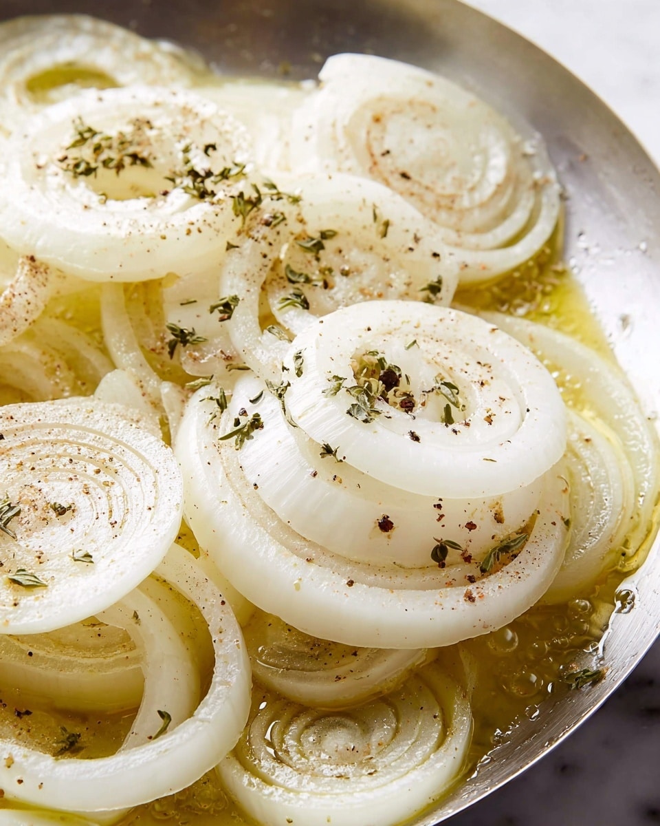 The image shows a close-up of several thin layers of white onion rings with some slices stacked in the center and others scattered around. The onions sit in a shiny metal pan where melted butter pools around the edges, giving a soft golden color. Black pepper and green herb flakes are lightly scattered on top of the onion layers. The texture of the onions looks fresh and slightly translucent, while the butter bubbles gently surround them. The scene is set on a white marbled surface. photo taken with an iphone --ar 4:5 --v 7