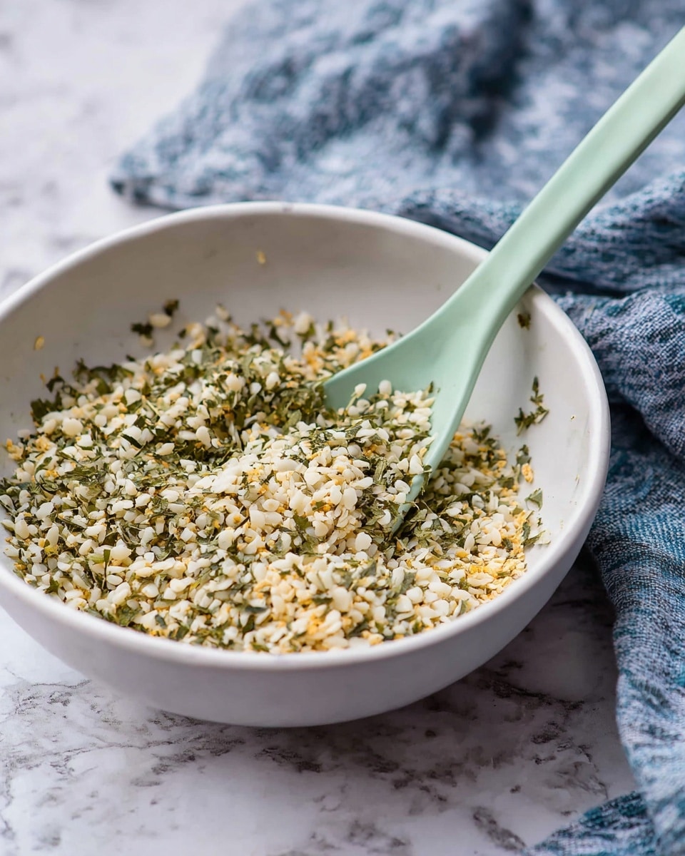 A white bowl filled with a mix of small white and light yellow grains, combined with green herb pieces, showing a fine, loose texture. A pale green spoon is dipped into the mixture, positioned on the right side of the bowl. The bowl sits on a white marbled surface, and a blue and white cloth is draped softly in the background. Photo taken with an iphone --ar 4:5 --v 7