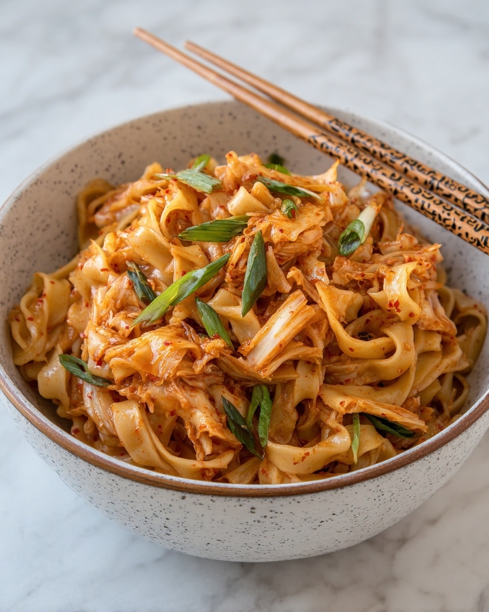 A bowl filled with thick, flat noodles coated in a light brown sauce or seasoning, mixed with pieces of reddish-orange kimchi scattered on top and throughout. Bright green sliced scallions are sprinkled over the dish in layers, adding a fresh pop of color. The noodles have a soft, slightly textured look, while the kimchi pieces show slight wilted edges. The bowl is white with a gray speckled pattern and a brown rim, sitting on a white marbled surface. A pair of light wooden chopsticks with a dark pattern lies on the bowl’s edge. Photo taken with an iphone --ar 4:5 --v 7