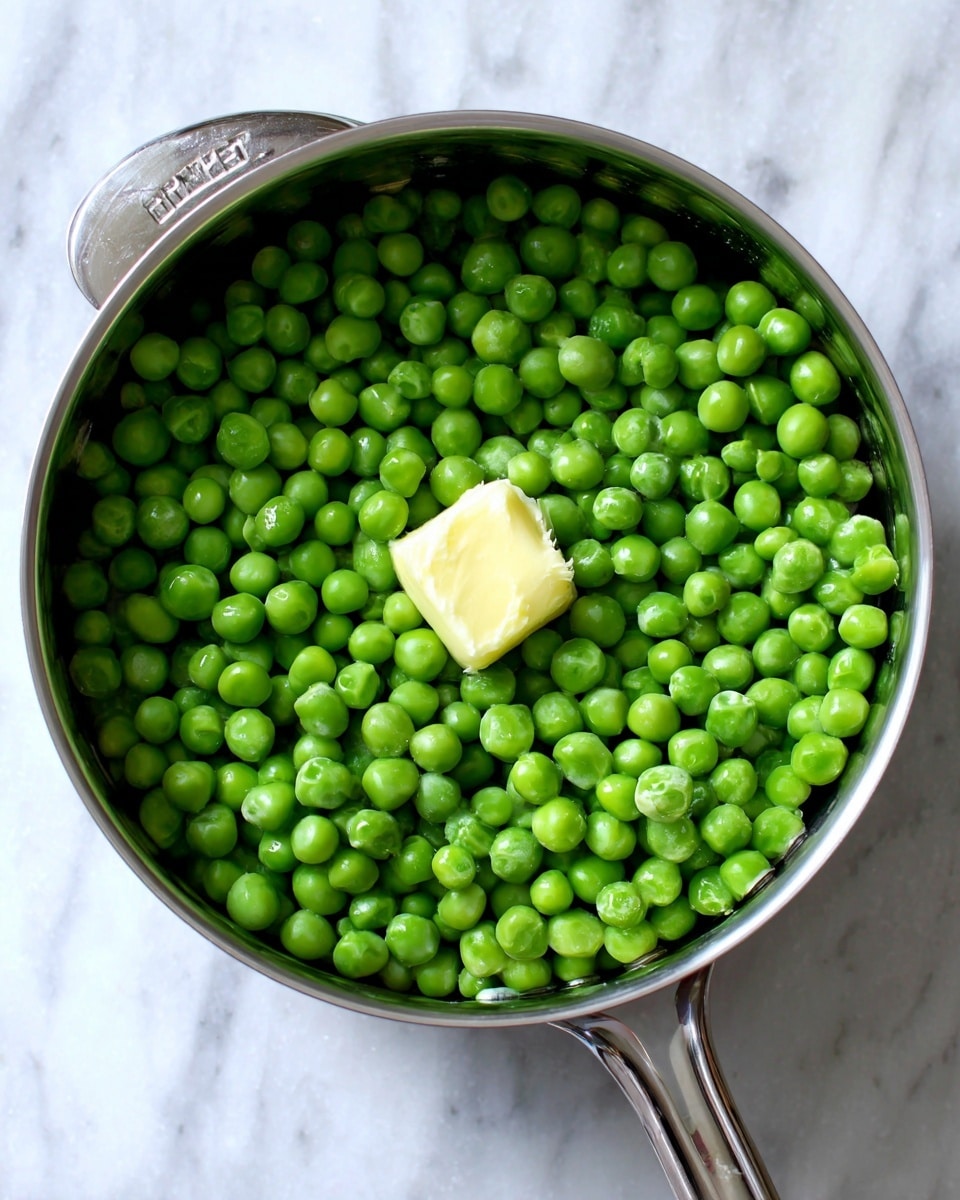 A shiny silver pot filled with bright green frozen peas that cover the entire bottom, with a small dollop of pale yellow butter resting in the center on top of the peas. The pot has measurement marks inside and smooth metal texture around the peas. The image shows the pot from above, placed on a white marbled surface. photo taken with an iphone --ar 4:5 --v 7