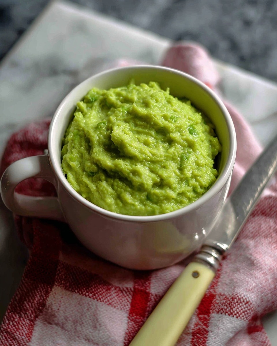 A close-up of a white cup filled with bright green mushy peas, showing a slightly chunky and soft texture on top. The cup sits on a red and white checkered cloth on a white marbled surface, with a silver-edged knife with a pale yellow handle lying next to it. The background is a blurred dark grey stone texture. photo taken with an iphone --ar 4:5 --v 7
