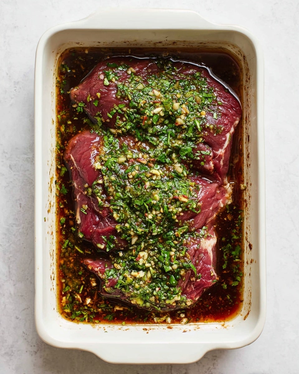 A rectangular white baking dish sits on a white marbled surface, holding two large pieces of raw dark red meat partially covered by a shiny brown marinade. The marinade is topped with a layer of finely chopped bright green herbs and minced garlic, spread evenly over the meat's surface, giving a fresh and textured look. The edges of the dish show darker marinade stains, adding contrast to the clean white dish. Photo taken with an iphone --ar 4:5 --v 7