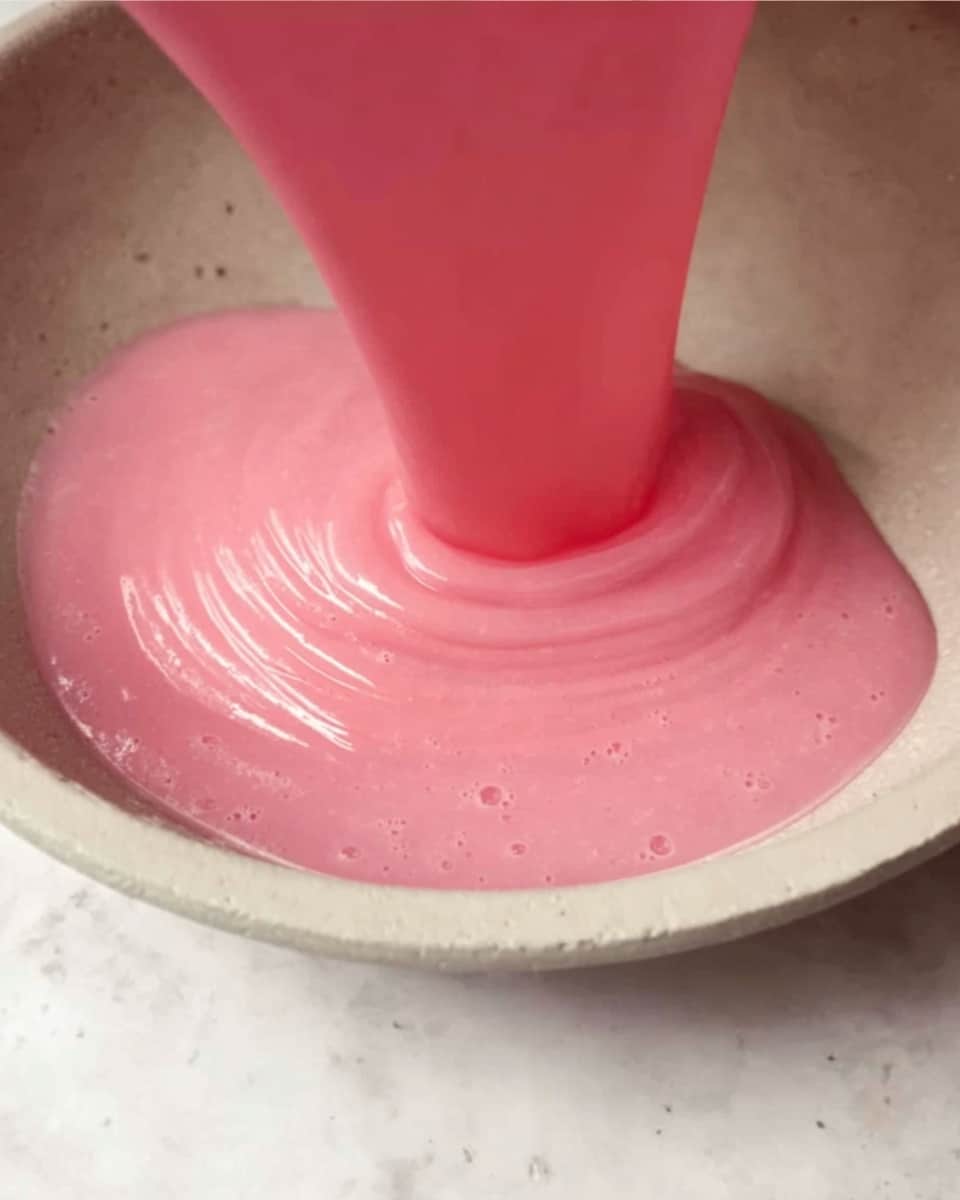 The image shows a smooth, thick pink batter being poured in a round, shallow white bowl with a slight matte texture. The pink batter has a glossy, silky surface and appears to be flowing gently, creating soft, curved layers as it spreads evenly across the base of the bowl. The background is a white marbled texture with subtle grey spots, giving a clean and bright contrast to the vibrant pink batter. photo taken with an iphone --ar 4:5 --v 7