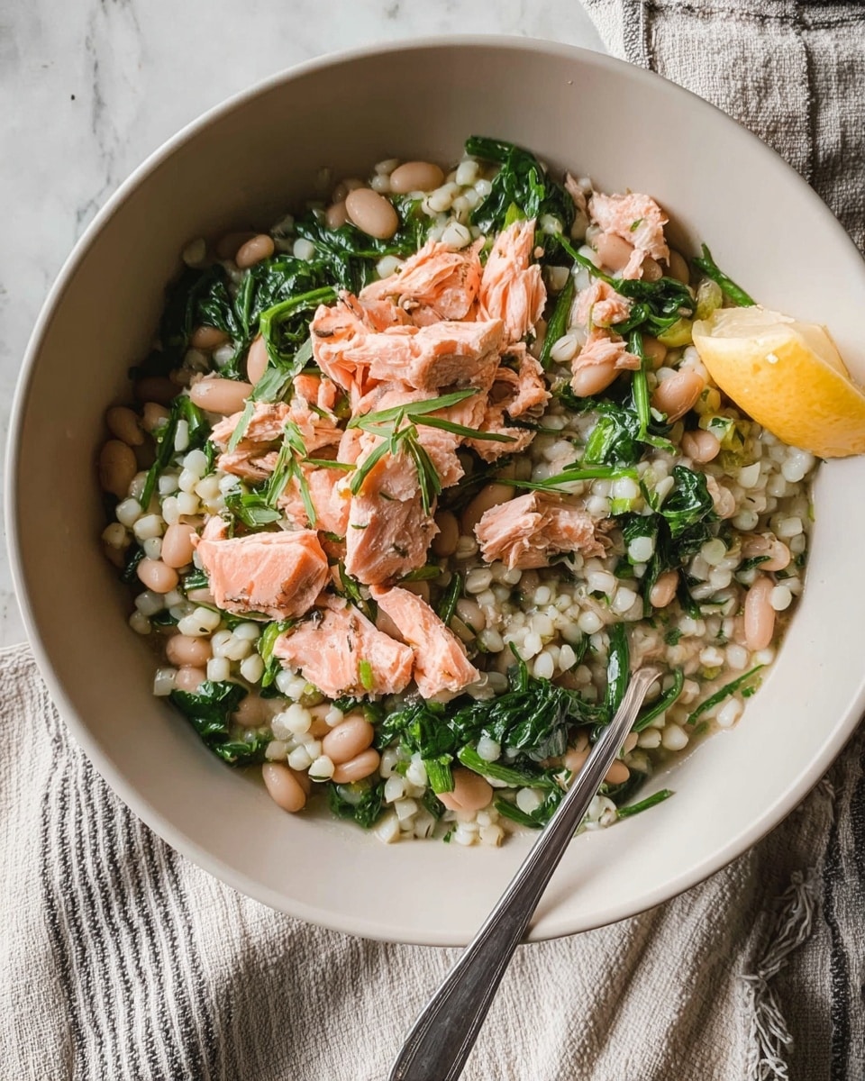 A white bowl filled with a dish made of three main layers: the bottom layer is a mix of white grains and white beans giving a soft, slightly bumpy texture; the middle layer has bright green leafy vegetables spread evenly throughout; the top layer is pieces of cooked pink fish flakes placed on top, some small green herb sprigs scattered for garnish, and a small slice of yellow lemon resting on the side. A silver spoon is partially inserted into the bowl from the right side. The bowl is placed on a white marbled surface with a striped cloth visible underneath. Photo taken with an iphone --ar 4:5 --v 7