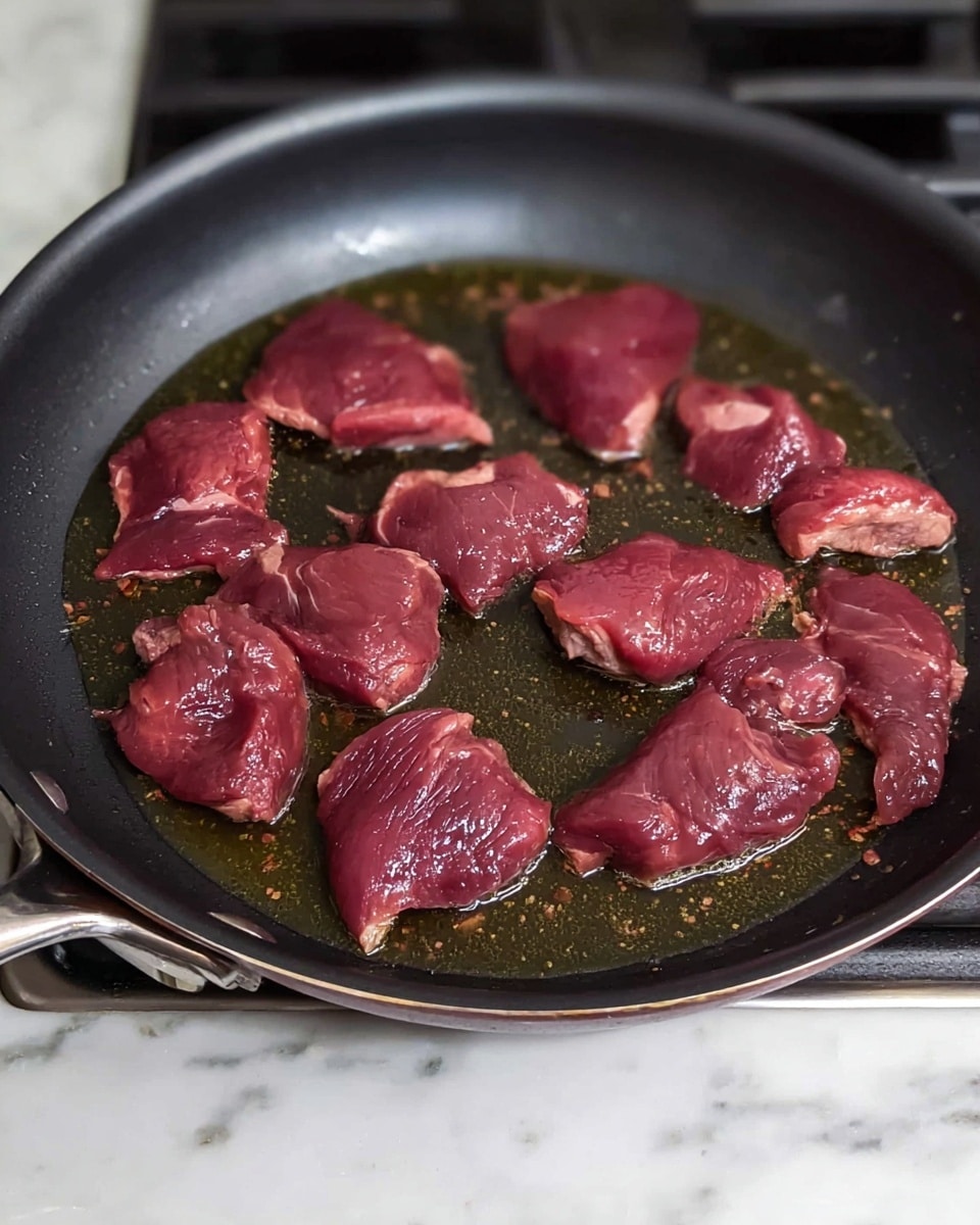 This image shows a dark non-stick frying pan on a stove with a white marbled countertop visible on the side. Inside the pan, several raw pieces of dark red meat with a smooth, shiny texture are frying in a layer of oil. The meat pieces are scattered but closely packed, each with uneven edges and some light spots of fat. The oil glistens around the meat, giving a wet, glossy look to the cooking surface. photo taken with an iphone --ar 4:5 --v 7