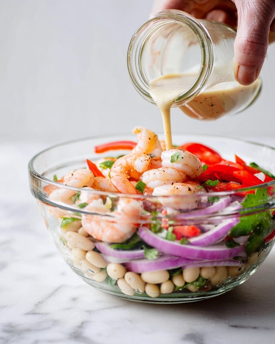 A clear glass bowl on a white marbled surface holds a colorful shrimp salad. The salad has four main layers: at the bottom are small white beans mixed with chopped herbs, above that bright red bell pepper strips, then light purple onion rings scattered around, and whole pink shrimp sitting on top. Fresh green herbs are mixed throughout. A woman's hand is pouring a smooth, light cream-colored dressing from a small glass jar over the salad. The scene is bright and clean with soft natural light, photo taken with an iphone --ar 4:5 --v 7
