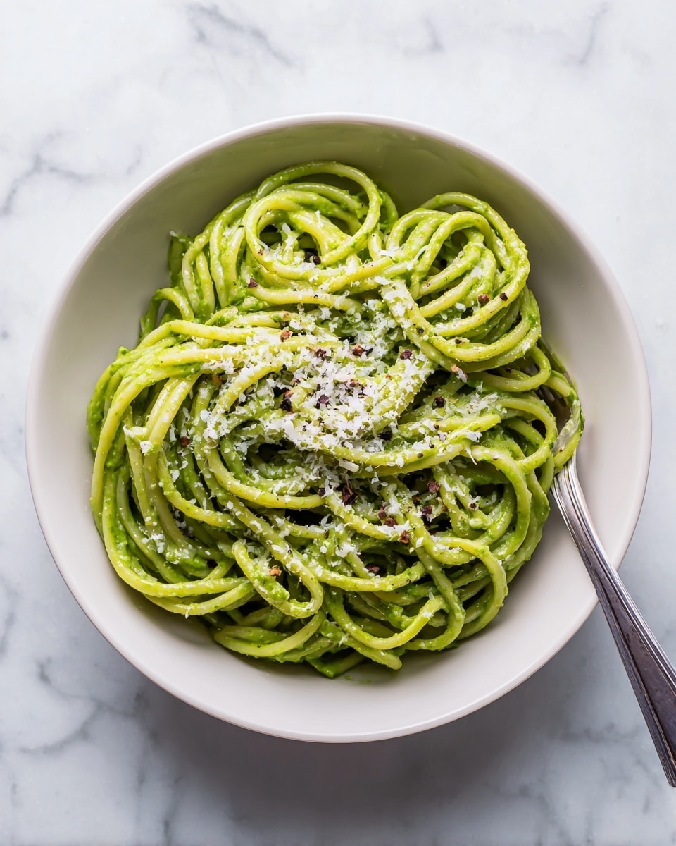 A white bowl holds a single layer of green pasta noodles coated in a smooth, vibrant green sauce. On top, there is a sprinkling of finely grated white cheese spread evenly, with small black pepper flakes scattered throughout. A silver fork is partially visible at the bottom right side of the bowl, nestled in the pasta. The bowl sits on a white marbled surface, giving a clean, bright look to the image. photo taken with an iphone --ar 4:5 --v 7