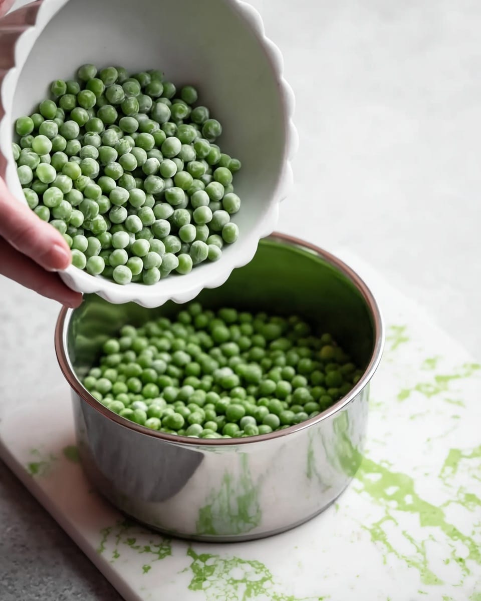 A close-up image shows a white bowl filled with small, round, green peas being tipped by a woman's hand over a shiny silver pot. The peas are bright and fresh, with a slightly matte texture, inside the bowl with scalloped edges. The pot is clean and reflective, placed on a white marbled surface that contrasts with faint green splashes of color on a white cutting board beneath. The composition focuses on the motion of the peas moving from the white bowl into the silver pot, creating a simple kitchen scene photo taken with an iphone --ar 4:5 --v 7