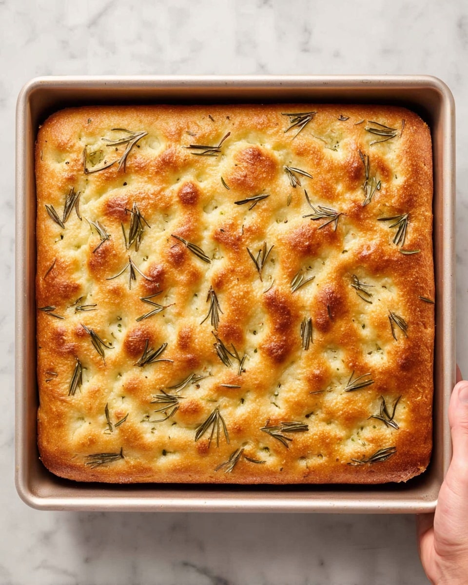The image shows a rectangular golden-brown focaccia bread baked in a rectangular pan with slightly raised edges. The top layer of bread is textured with an uneven surface, dimpled spots, and is dotted with scattered green rosemary leaves. The bread edges are slightly darker, indicating a crisp crust, while the middle appears soft and fluffy. The pan is being held by a woman's hand on the right side, and the whole setup sits on a white marbled surface. photo taken with an iphone --ar 4:5 --v 7