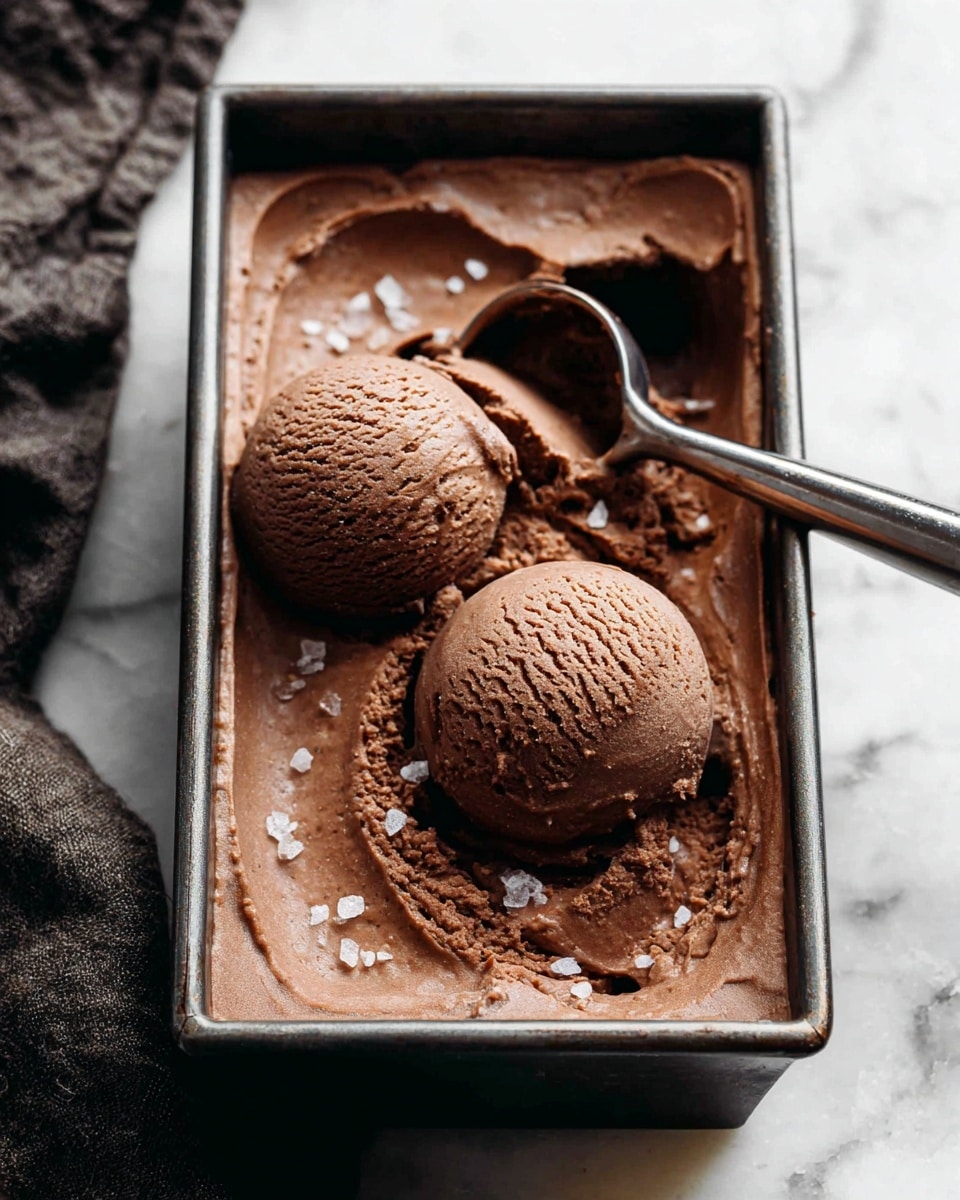 Two round scoops of chocolate ice cream, rich and creamy with a smooth slightly melty texture, sit side by side in a rectangular metal tray filled with more chocolate ice cream spread evenly across the base. A metal ice cream scoop rests in the tray next to the scoops. The ice cream has small specks of salt sprinkled on top. The tray is set on a white marbled surface. Photo taken with an iphone --ar 4:5 --v 7