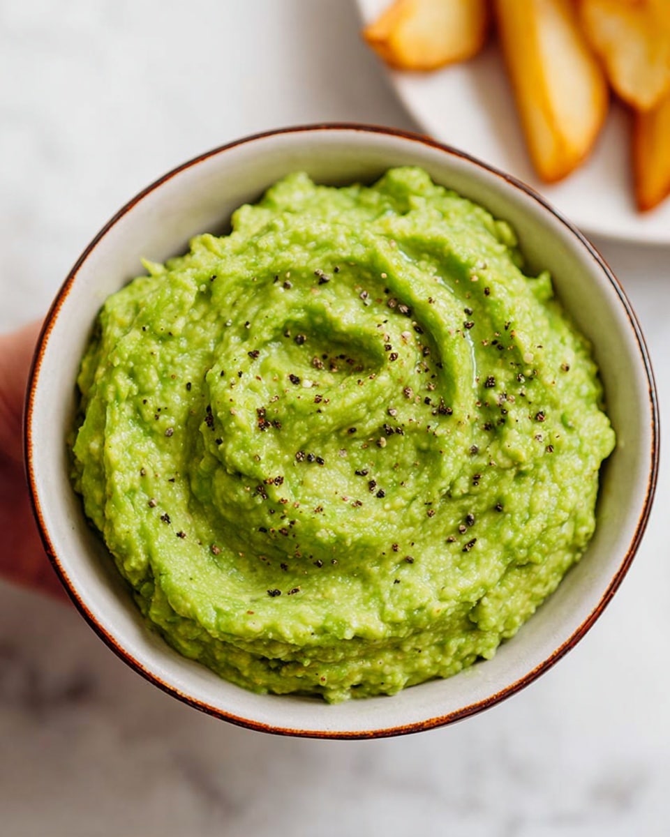 A bowl filled with bright green guacamole, showing a slightly chunky texture with small pieces of avocado visible. The guacamole has a swirled pattern on the surface and is speckled with cracked black pepper. The bowl is white with a thin brown rim, placed on a white marbled surface. In the background, a woman's hand is holding a white plate with light yellow potato wedges that are partially blurred. Photo taken with an iphone --ar 4:5 --v 7