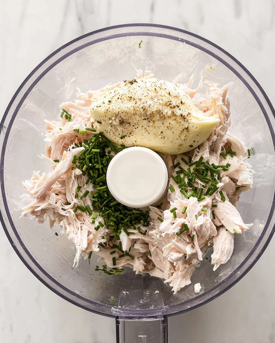 Inside a clear food processor bowl, there are several layers of ingredients visible from above. The bottom layer consists of shredded pale pink cooked chicken pieces, which are tender and fibrous. On top of the chicken toward the center is a smooth, creamy dollop of pale yellow mayonnaise, sprinkled lightly with black pepper. Scattered over the chicken and mayonnaise are small chopped green herbs, likely chives, adding a fresh vibrant contrast to the soft colors. The food processor's central blade has a white plastic cover. The bowl sits on a white marbled surface. Photo taken with an iphone --ar 4:5 --v 7
