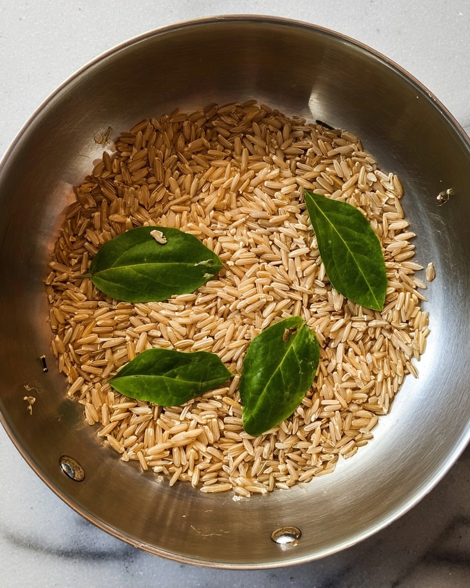 A shiny metal pan filled with light brown grains that look like toasted rice, scattered unevenly across the bottom. Among the grains are four fresh green leaves, two larger and two smaller, placed on top of the grains, adding a pop of color. The pan has a smooth, reflective surface with a small metal rivet visible near the edge, and the background is a white marbled texture. Photo taken with an iphone --ar 4:5 --v 7