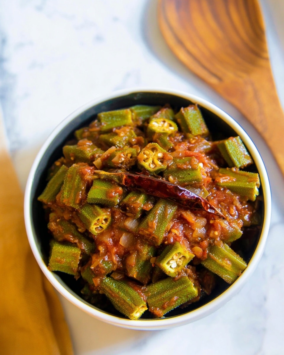 A close-up view of a bowl filled with a chunky okra dish cooked in a reddish-brown tomato-based sauce. The okra pieces are cut into thick, green segments mixed with the sauce that has visible tomato chunks and some white okra seeds. A single dried red chili is placed on top, giving a bit of color contrast. The food is served in a white bowl set on a surface with a white marbled texture, with a wooden spoon slightly blurred in the background. The overall image has bright natural lighting and warm tones. photo taken with an iphone --ar 4:5 --v 7