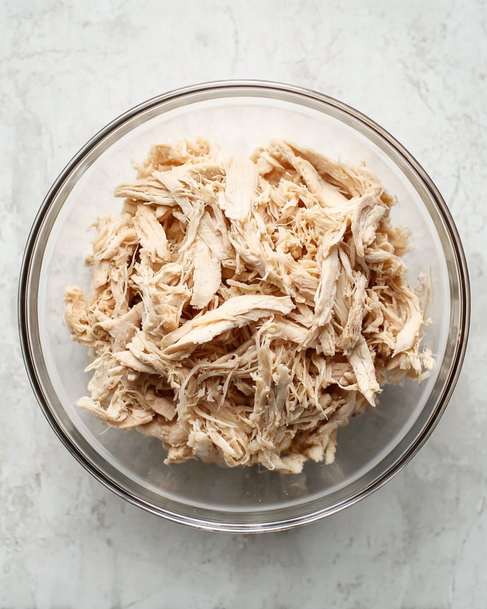 A clear glass bowl filled with shredded cooked chicken, showing many pale beige and light brown pieces loosely piled together. The chicken has a fibrous texture and is arranged evenly inside the bowl, which sits on a white marbled surface. The image is shot from above, focusing on the simple presentation of the chicken without any other items around photo taken with an iphone --ar 4:5 --v 7