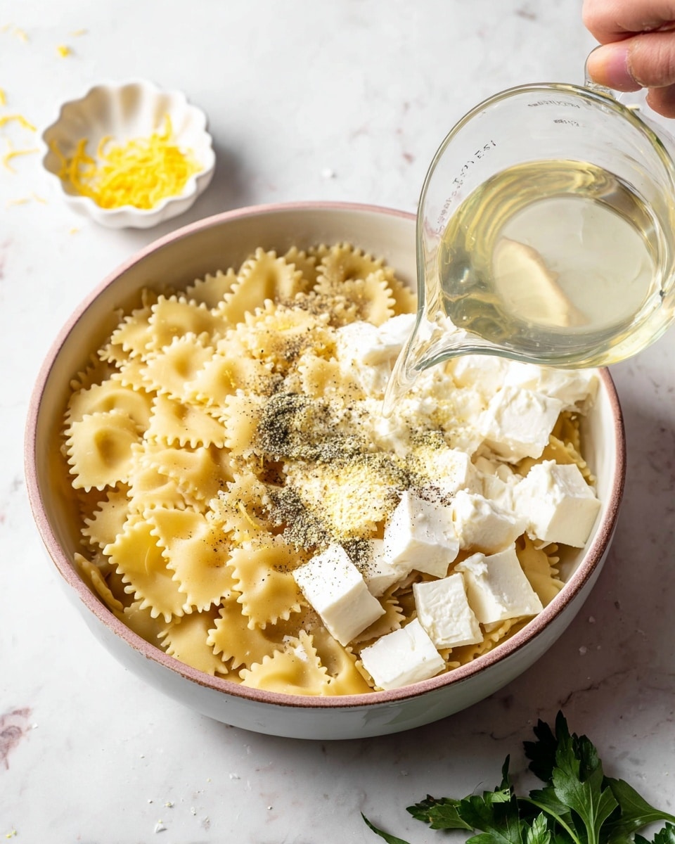A white bowl filled with short, square pasta with ruffled edges forms the base layer. On top of the pasta, there are several white cubes of soft cheese clustered on the lower right side. Sprinkled over the pasta and cheese are patches of ground black pepper and granulated yellow seasoning. A woman's hand is pouring a light, clear liquid from a glass measuring cup into the bowl from the lower left corner of the image. In the upper left, a small white dish holds scattered lemon zest, while fresh green parsley leaves rest partially visible at the bottom edge. The setting is on a white marbled surface. photo taken with an iphone --ar 4:5 --v 7